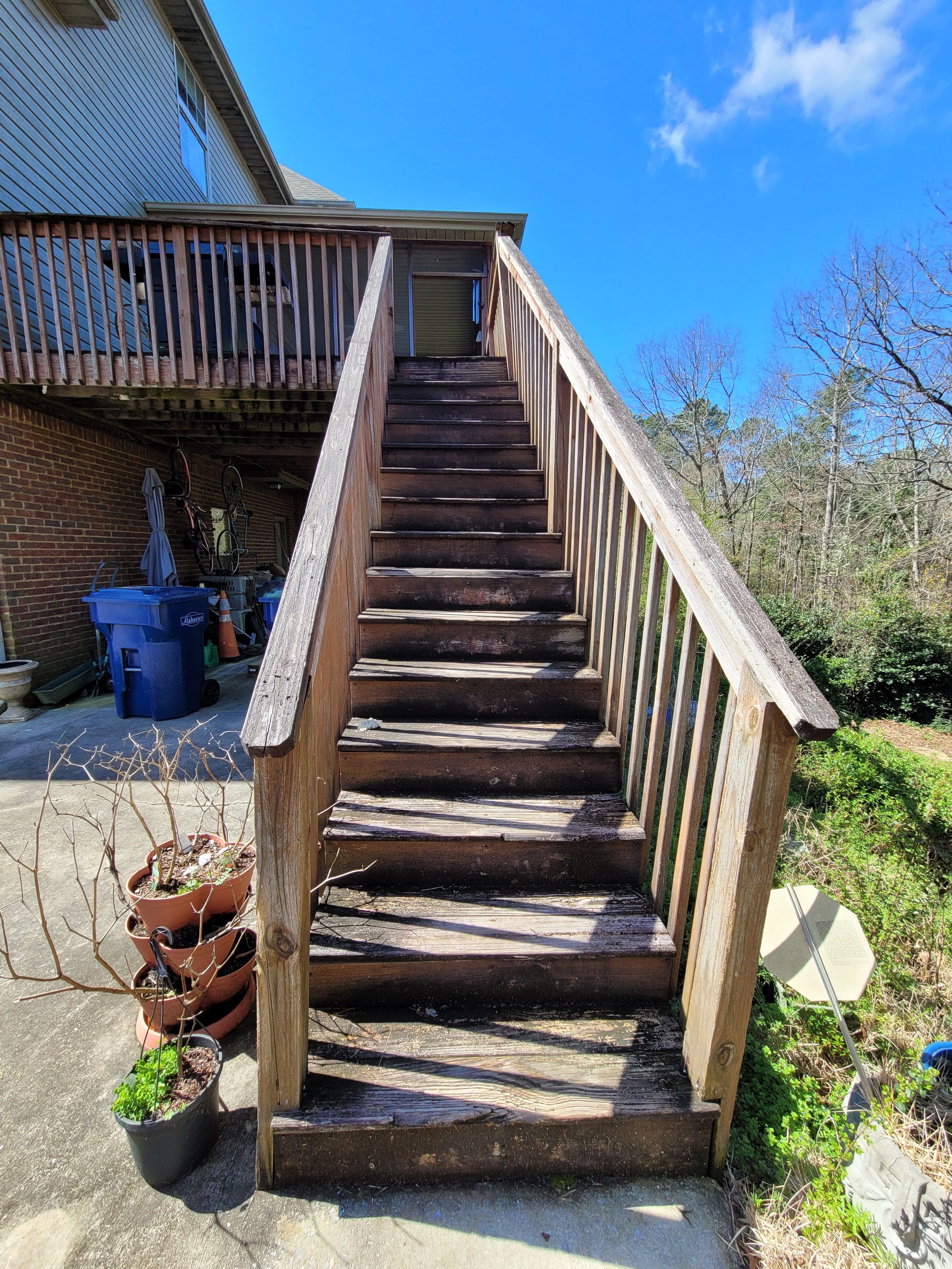 Wooden stairs leading to a deck, surrounded by trees and potted plants under a blue sky.