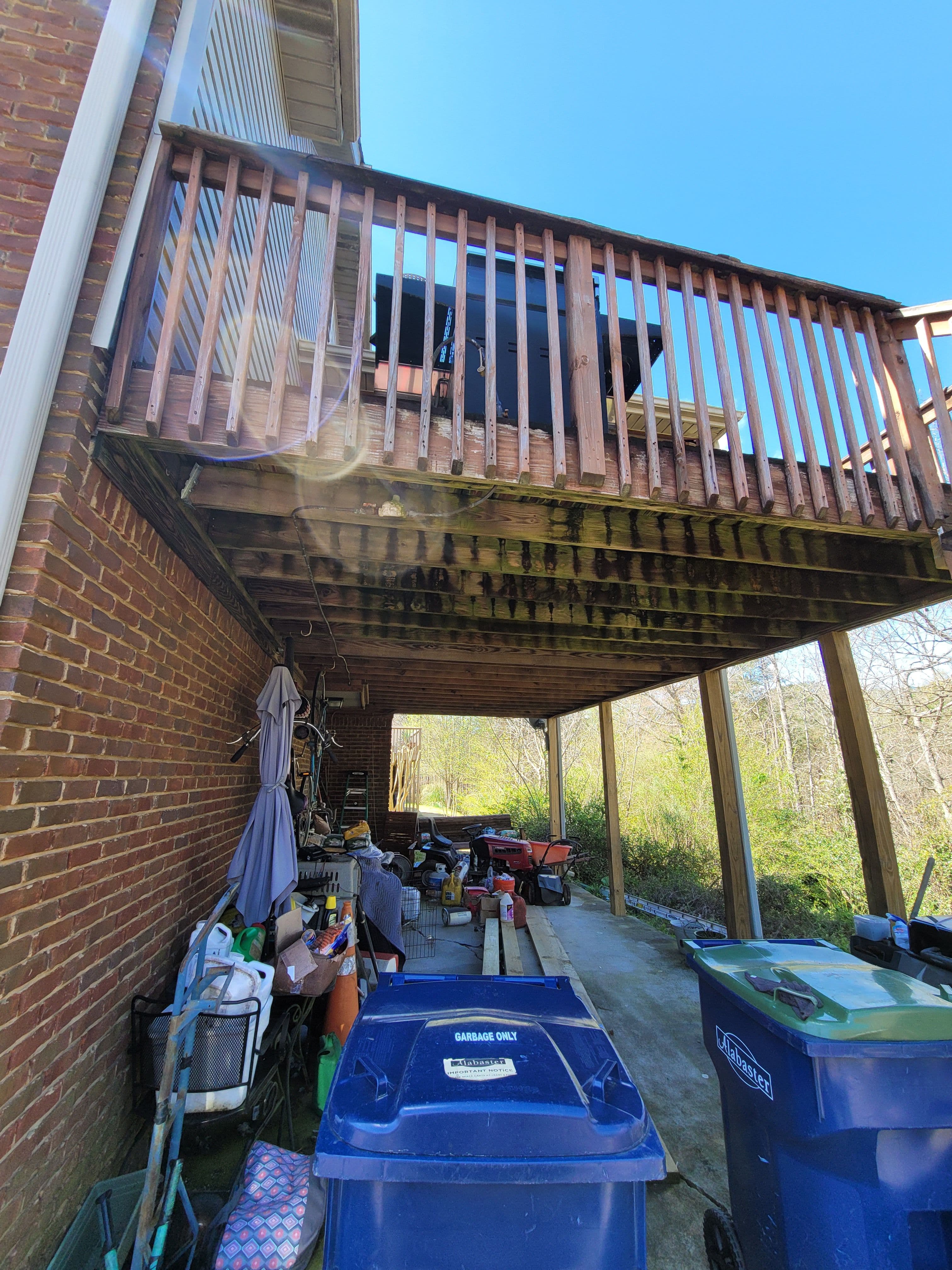 Underneath a wooden deck, cluttered with storage items and trash bins. Clear blue sky.