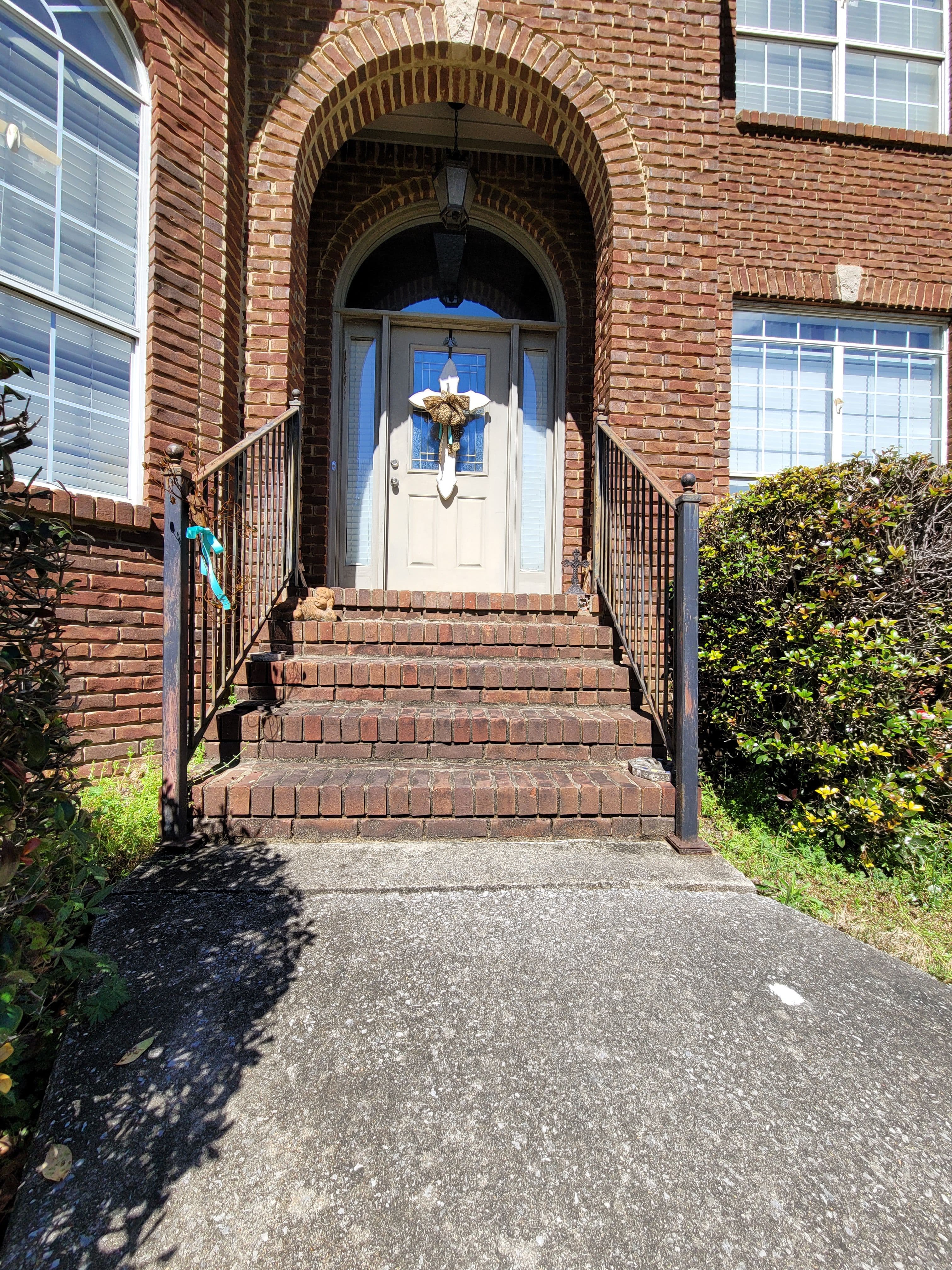 Brick entrance with steps, lantern, and a decorative wreath on the door. Lush greenery nearby.