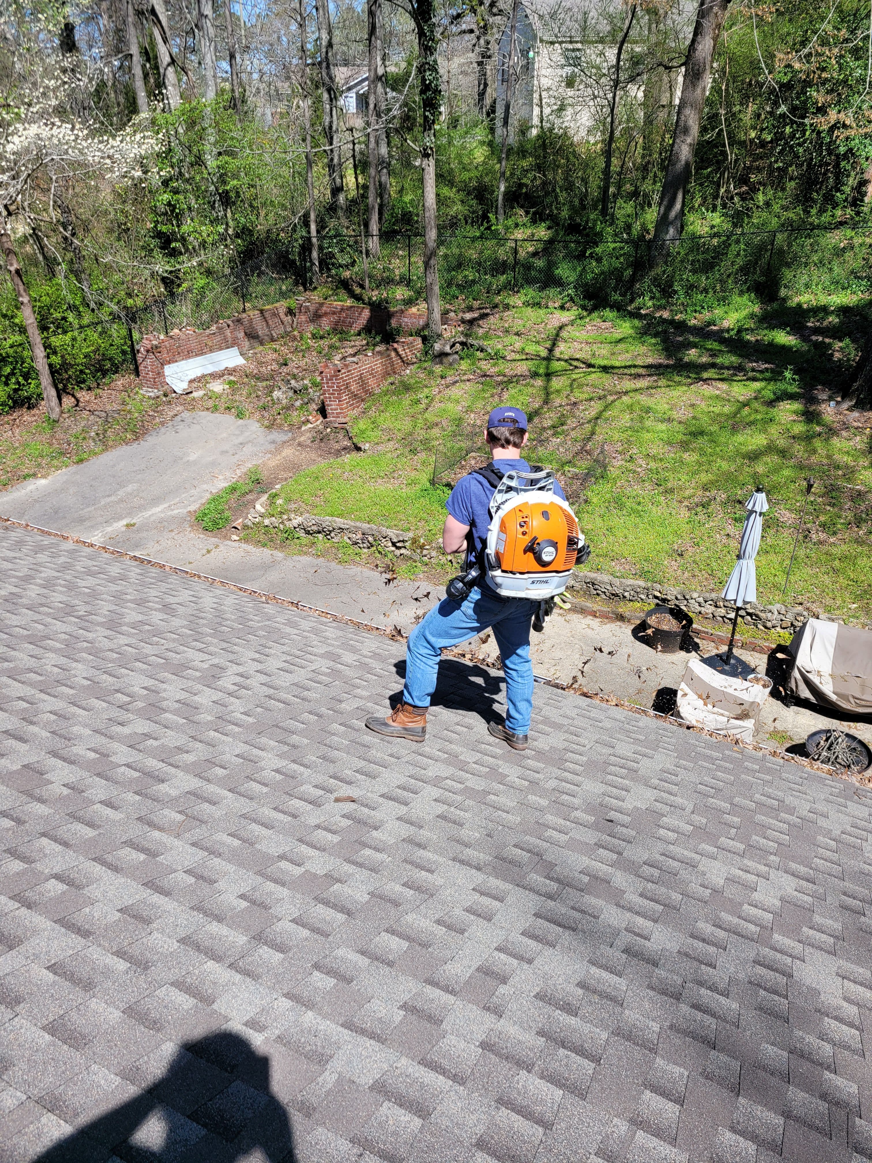 Person using a backpack blower on a roof with greenery and a driveway in the background.