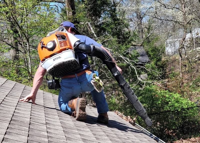 A Do-Gooders worker kneeling on a shingle roof, clearing leaves and debris with a backpack blower