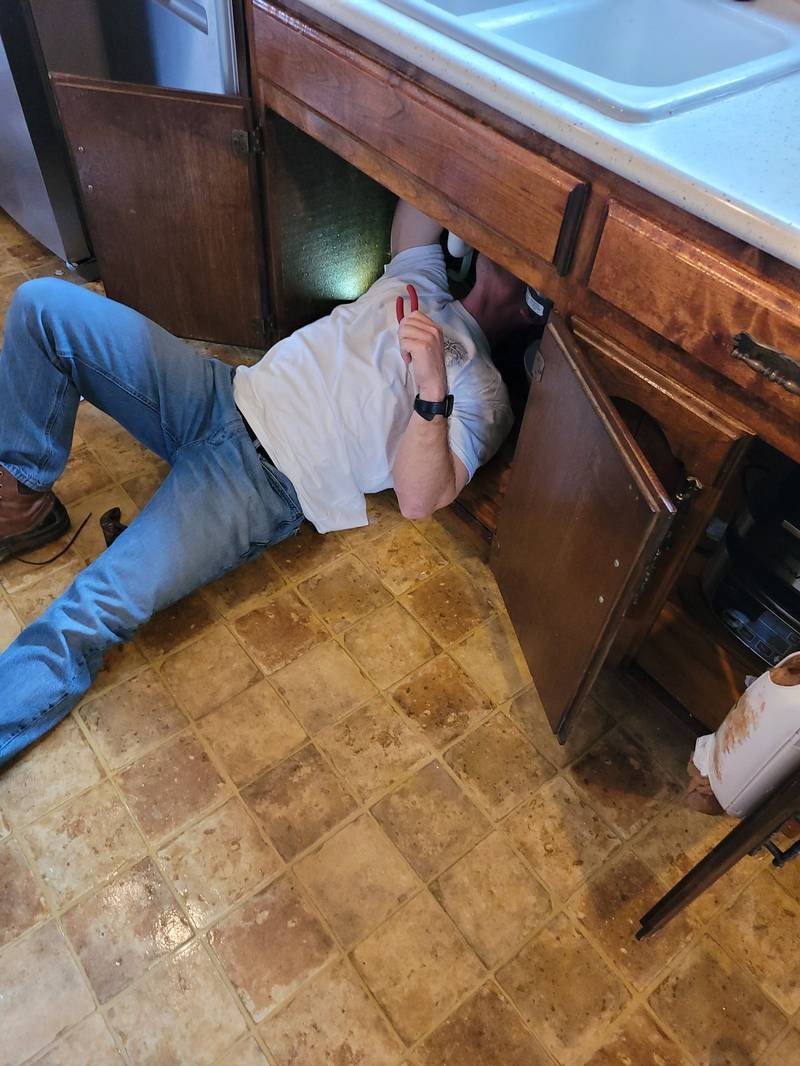 A worker lying on a kitchen floor reaching into the cabinet under the sink to make a repair